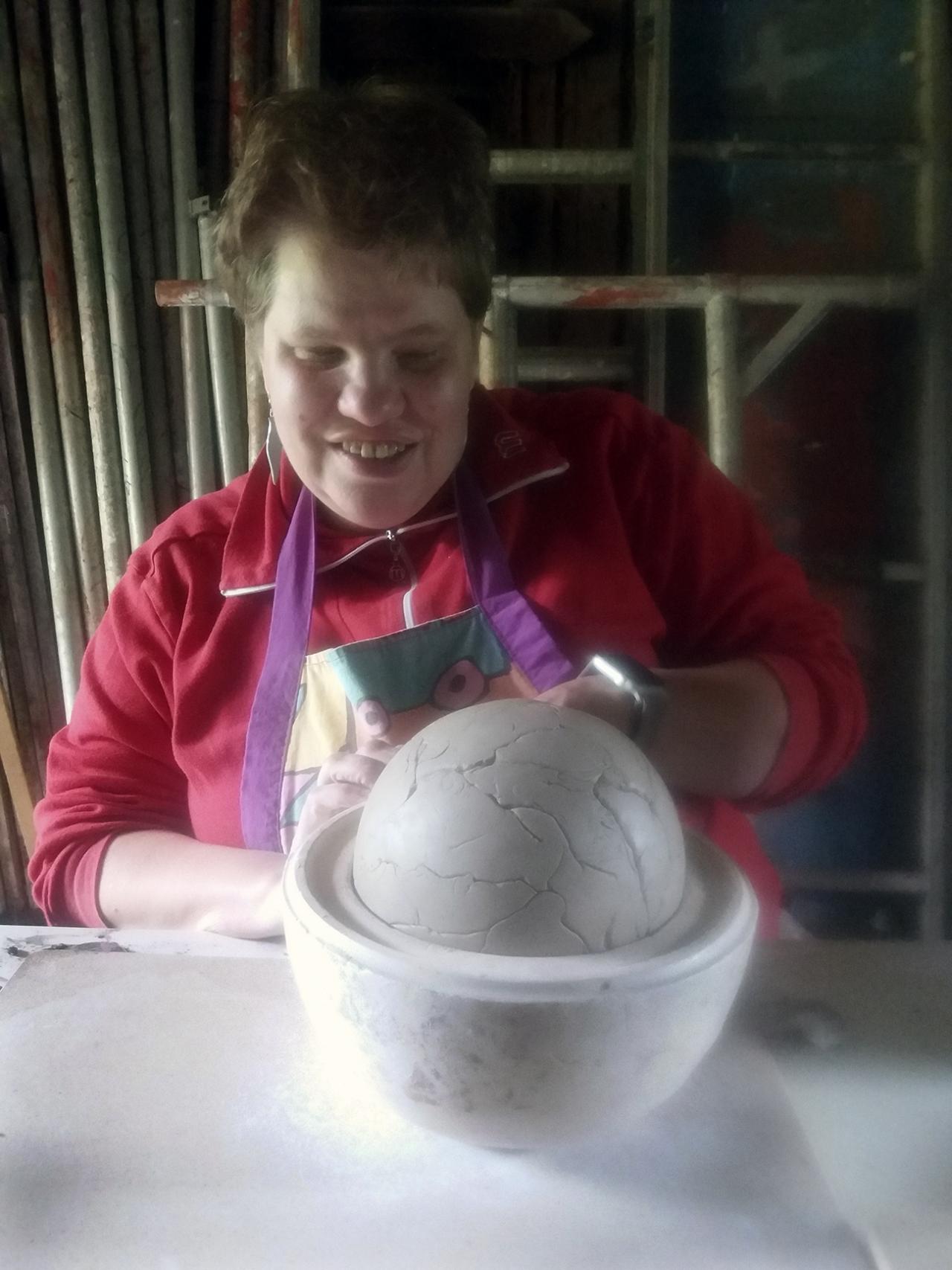 Heidi työstää savesta keraamista teepannua puolipallomuotin avulla. Heidi hymyilee ja hänellä on essu päällä. 